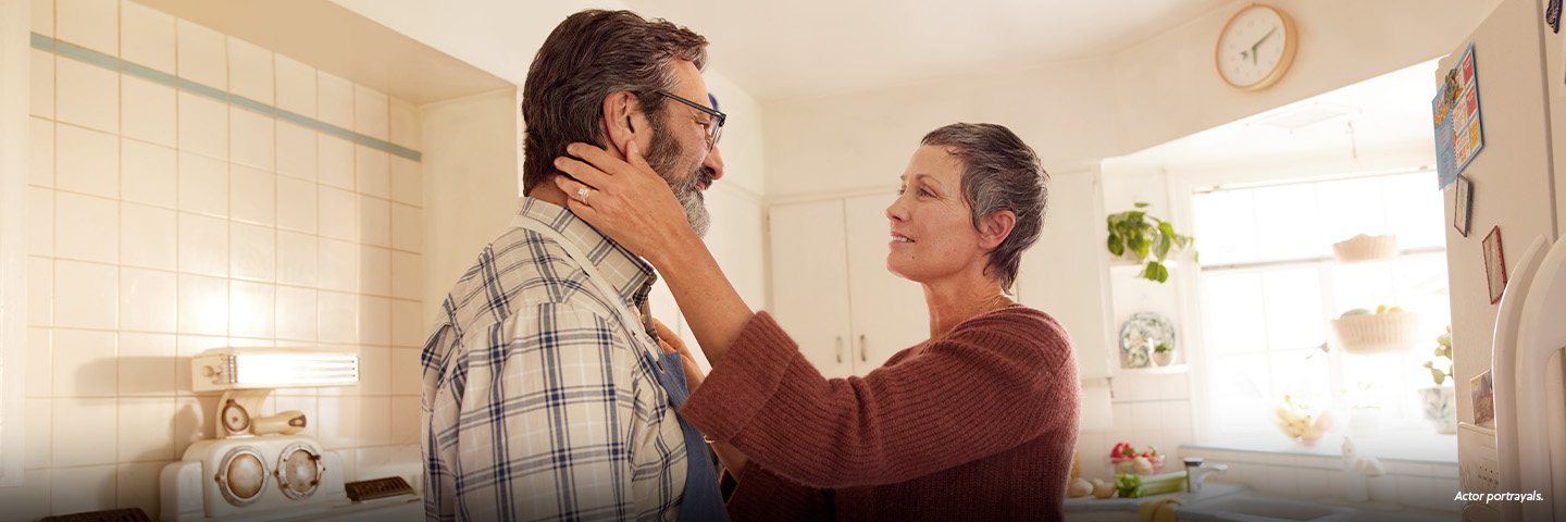 A Latina woman in her 50s with short gray hair is standing in the kitchen with her partner, a Latino man in his 50s. The woman is reaching out, with one hand resting on the man's shoulder and the other hand gently holding the man's jaw. They are looking at each other fondly. Not actual patients.