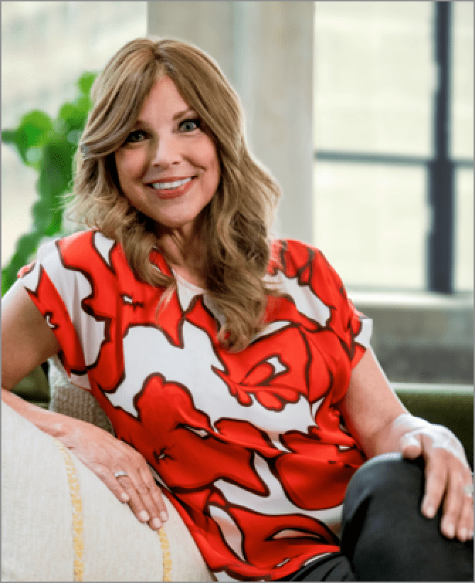 Jacquie, a woman receiving TRODELVY, is sitting on a green couch with a white pillow while smiling at the camera. Jacquie is a White woman wearing a brown wig with blonde highlights, a shirt with a red and white pattern, and black pants.