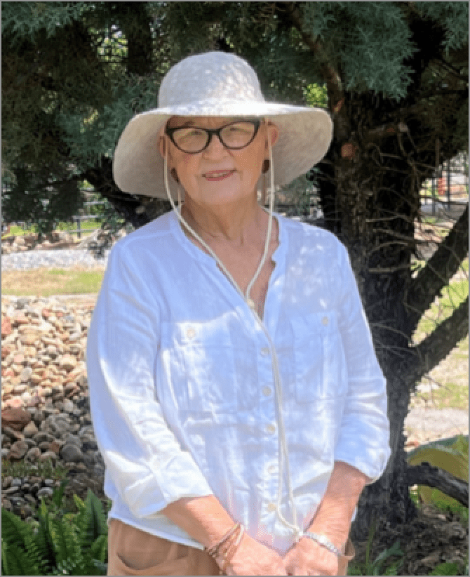 Jana, a woman receiving TRODELVY, stands outdoors smiling at the camera. Jana is a White woman wearing a white sunhat, glasses, and a white linen shirt.