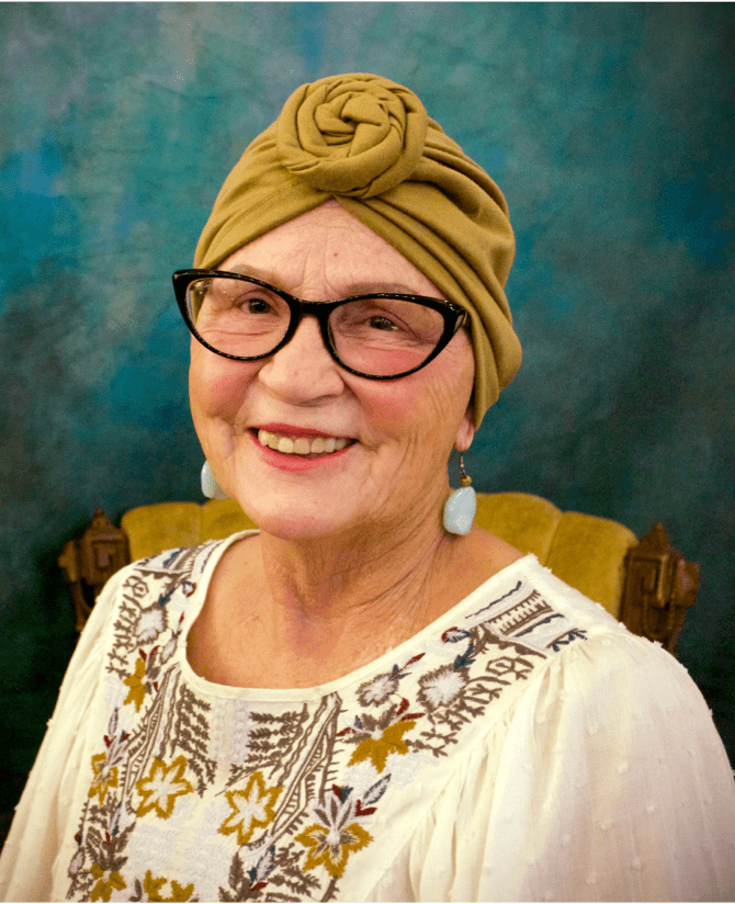Jana, a woman receiving TRODELVY, sits smiling at the camera. Jana is a White woman wearing a headscarf, glasses, and a white patterned shirt.