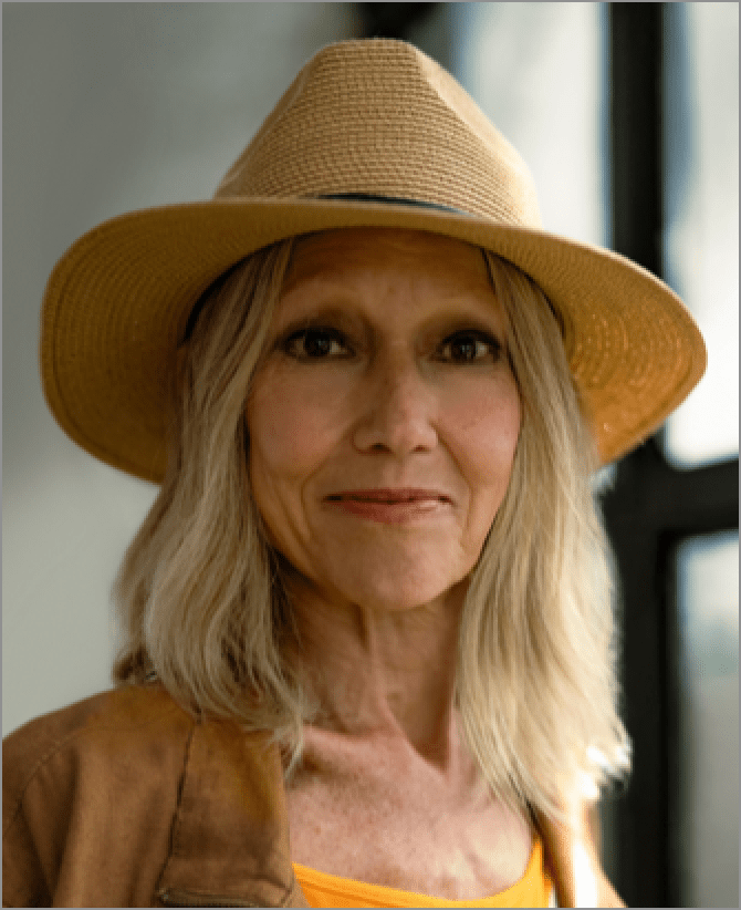 Karen, a woman receiving TRODELVY. Karen is a White woman wearing a blonde wig, a brown sun hat, a brown leather jacket, and a white shirt.