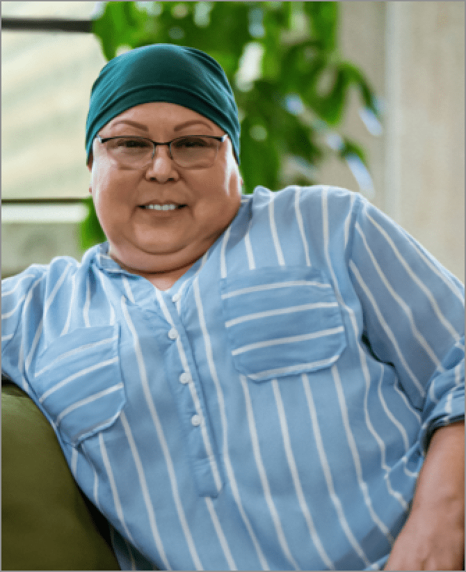 Lisa, a woman receiving TRODELVY, sits smiling at the camera. Lisa is a Latina woman wearing a headscarf, glasses, and a blue shirt with white stripes.