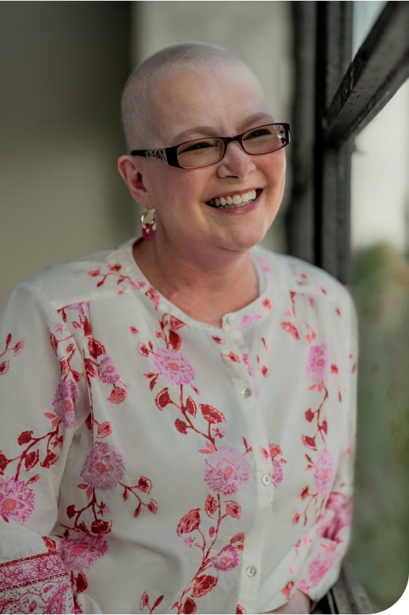 Tanya, a woman receiving TRODELVY, sits smiling at the camera. Tanya is a White woman with no hair, wearing glasses and a white shirt with a red floral pattern.