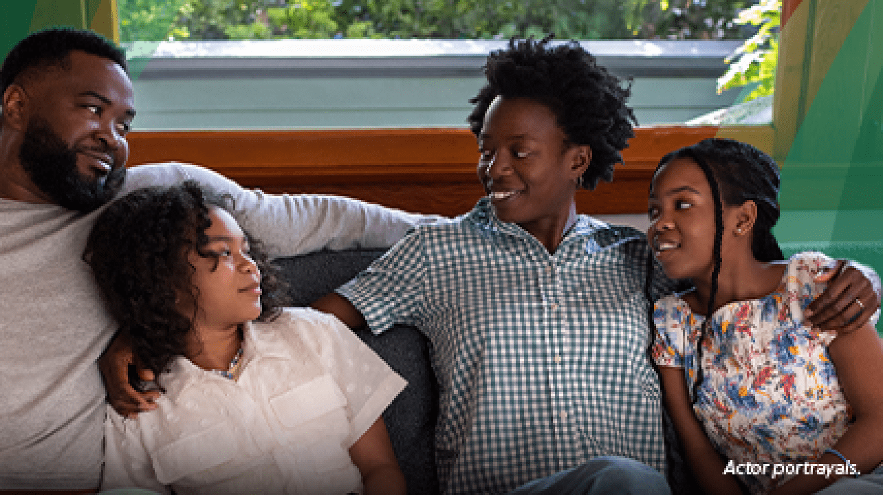 A Black woman in her mid-40s with short natural hair is cuddled on the living room couch with her family--a Black man in his 40s and their 2 preteen daughters. The woman has her arms around both of her daughters and is smiling at her husband. The family is close, leaning on each other and smiling. Actor portrayals.