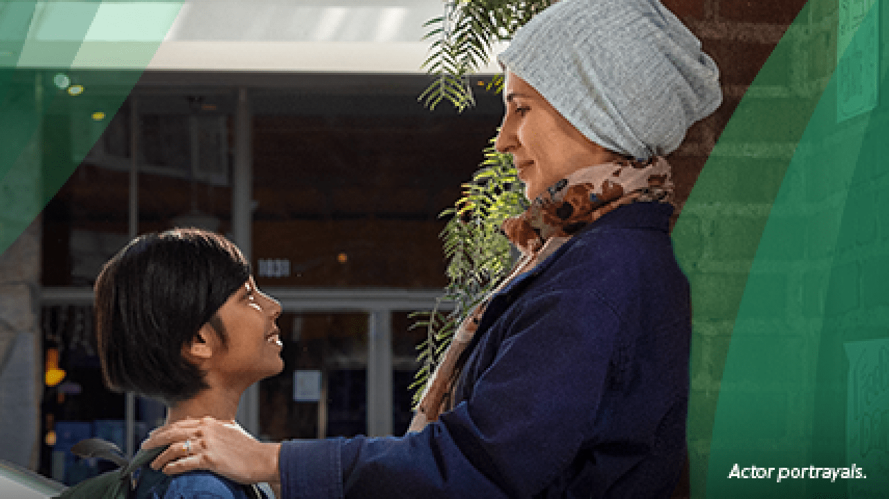 A Latina woman in her 40s wearing a light blue knit hat is dropping her 10-year-old son off at school. They are standing in the doorway facing each other, and the woman has her hands on the boy's shoulders. Actor portrayals.